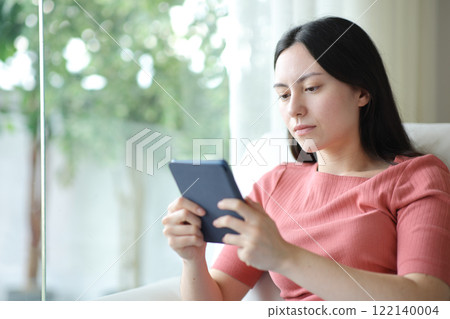 Serious asian woman reading ebook on a sofa 122140004