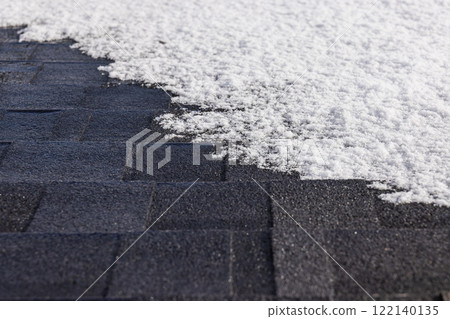 Snow-covered asphalt shingle roof with visible patterns and texture, illuminated by winter sunlight creating a striking contrast 122140135