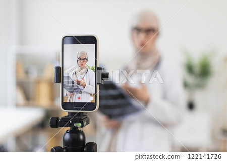 Muslim woman doctor creating educational video with x-ray for online audience. Adult female in hijab sharing medical knowledge through smartphone camera. Muslim woman doctor creating educational video with x-ray for online audience. Adult female in hijab sharing medical knowledge through smartphone camera. 122141726