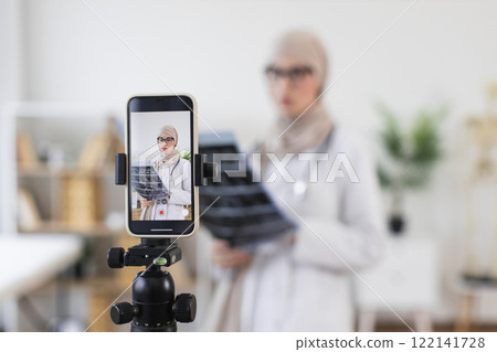 Young Muslim female doctor with x-ray recording medical video lesson on smartphone in modern office. Professional education focus. 122141728