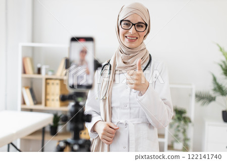 Young Muslim woman doctor records video lesson about healing techniques using smartphone. Smiling female in hijab and lab coat gives thumbs up, creating engaging content for online audience. Young Muslim woman doctor records video lesson about healing techniques using smartphone. Smiling female in hijab and lab coat gives thumbs up, creating engaging content for online audience. 122141740