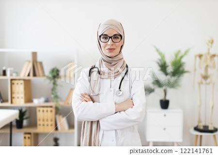 Portrait of confident young Muslim woman doctor in professional office. She wears glasses, stethoscope, and hijab, projecting professionalism and expertise. Background features office decor and plants 122141761