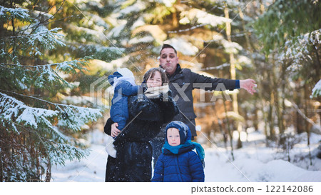 Happy family enjoying a winter day, playing joyfully in the snow amidst a beautiful forest filled with trees and sunlight 122142086