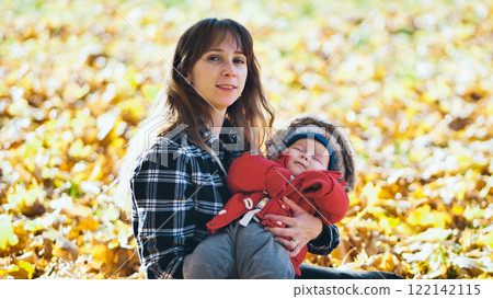 Young mother holding her sleeping baby in a beautiful autumn park, surrounded by colorful fallen leaves Young mother holding her sleeping baby in a beautiful autumn park, surrounded by colorful fallen leaves 122142115