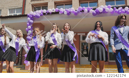 Joyful graduates in purple ribbons joyfully mark their last day of school, exchanging gestures and blowing air kisses outside a school adorned with balloons Joyful graduates in purple ribbons joyfully mark their last day of school, exchanging gestures and blowing air kisses outside a school adorned with balloons 122142174