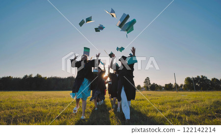 Happy graduates tossing their books in the air and running through a field at sunset 122142214