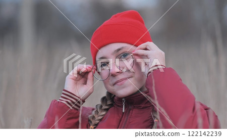Teenage girl with braids, red beanie, and round glasses, standing in grassy field and touching eyewear with confident smile 122142239