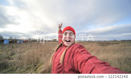 Cheerful female with braided hair in crimson winter outfit capturing selfie near colorful cottages amid barren landscape 122142248