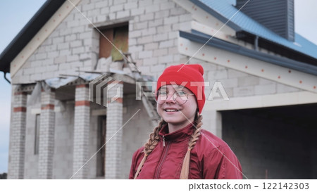 Confident young woman architect wearing a red beanie is smiling near a building under construction 122142303