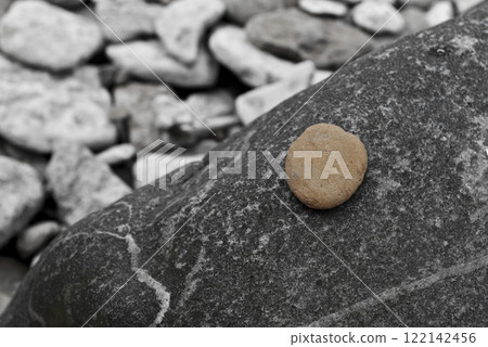 Simple composition featuring single light brown pebble resting on textured dark stone with blurred background of scattered rocks 122142456