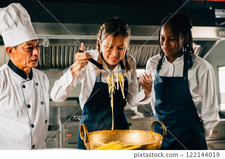 Chef coaches in kitchen. Schoolgirls craft Japanese noodle. Kids and teacher at stove. Smiling portrait of learning is modern education. Making dinner with ladle gives joy. 122144019