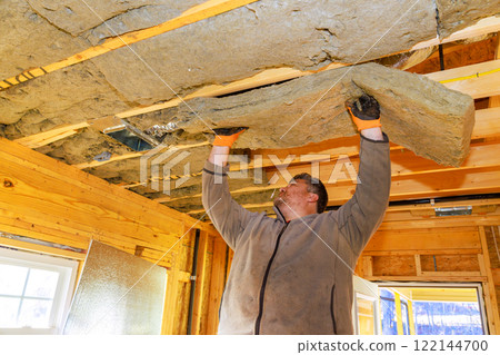 Carpenter wearing gloves raises insulation panels to fit them into wooden ceiling structure in renovations house. 122144700