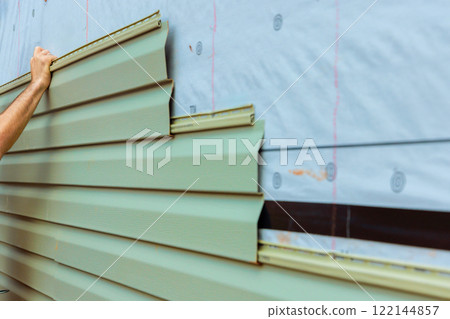 Construction worker carefully lays green siding on building exterior while ensuring proper fit. 122144857
