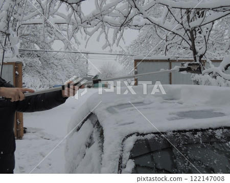 partially visible man in dark clothes removes layer of snow from roof of light colored car in private yard after heavy snowfall with long device, snow removal from car after winter storm partially visible man in dark clothes removes layer of snow from roof of light colored car in private yard after heavy snowfall with long device, snow removal from car after winter storm 122147008