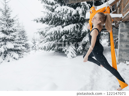 Young woman doing fly yoga in snowy forest in mountains. Yoga in hammock in winter against snow and fir trees. Gymnastic tricks outdoors in extreme conditions 122147071