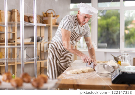 Elderly experienced male baker using knife to cut raw dough to make croissants 122147338