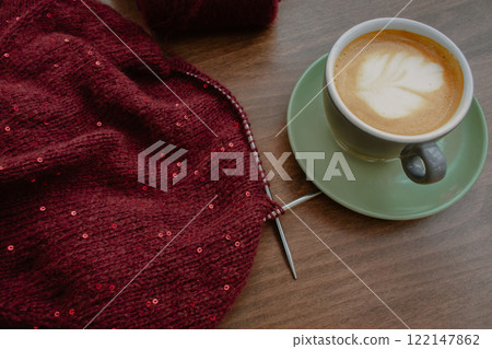 Red knitted sweater with sequins and knitting needles with coffee cup on table. Sequin pattern in needlework with cappuccino. Knitting hobby. Knitted fashion concept. Sequins pattern in knitwear. Red knitted sweater with sequins and knitting needles with coffee cup on table. Sequin pattern in needlework with cappuccino. Knitting hobby. Knitted fashion concept. Sequins pattern in knitwear. 122147862