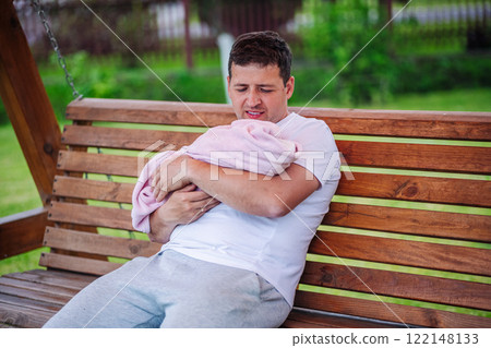 Caring father holding newborn daughter in soft pink blanket, sitting on wooden swing during peaceful backyard moment 122148133