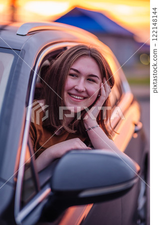 Smiling female driver relaxing inside vehicle, golden sunlight illuminating interior during scenic sunset drive 122148144