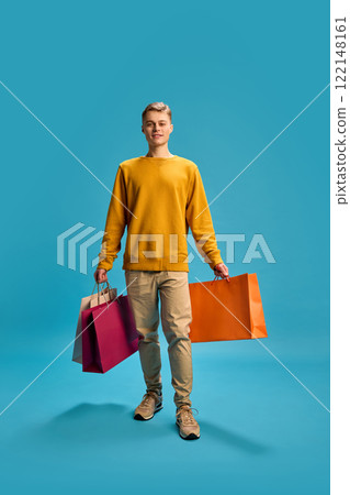 Full-length photo of young handsome man dressed casual outfit, walking with shopping bags against blue studio background. Full-length photo of young handsome man dressed casual outfit, walking with shopping bags against blue studio background. 122148161