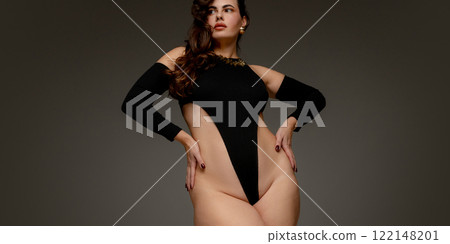 Plus size woman standing tall in black bodysuit, holding hands on hips, radiating strength, confidence, and natural elegance against grey studio background. 122148201