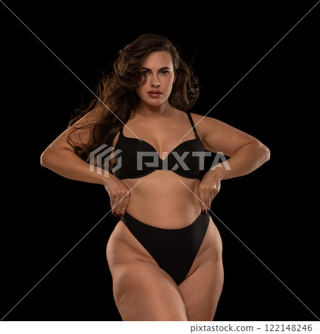 Confident woman, model XXL, in black lingerie, posing holding hands on waist, celebrating strength and individuality against black studio background. 122148246
