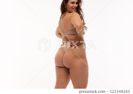 Back view young confident woman, plus size model, posing in beige lingerie, looking at camera over shoulder against white studio background. Back view young confident woman, plus size model, posing in beige lingerie, looking at camera over shoulder against white studio background. 122148283