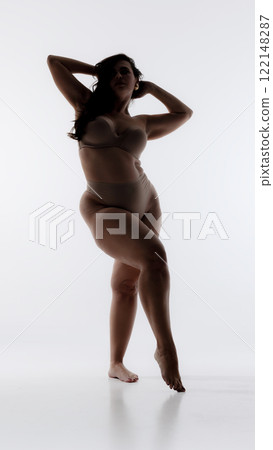 Silhouette of woman standing gracefully in lingerie, arms raised behind head, exuding elegance and empowerment against white studio background. 122148287