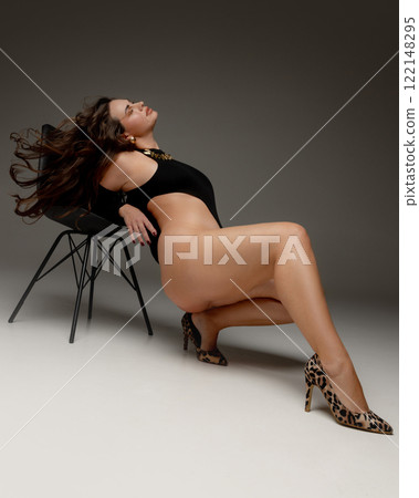Woman, model plus size, leaning back on chair, her hair flowing freely, celebrating empowerment and femininity against grey studio background. Woman, model plus size, leaning back on chair, her hair flowing freely, celebrating empowerment and femininity against grey studio background. 122148295