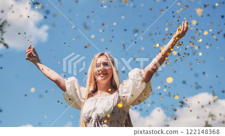 Young blonde woman throwing golden confetti in the air, celebrating success with arms raised and a cheerful smile under a blue sky Young blonde woman throwing golden confetti in the air, celebrating success with arms raised and a cheerful smile under a blue sky 122148368
