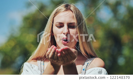 Blonde woman softly blowing kiss, lush green background, sunlight warming her face 122148370