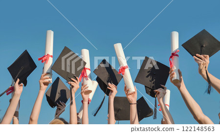 Students celebrating graduation by raising their diplomas and mortarboards against a clear blue sky Students celebrating graduation by raising their diplomas and mortarboards against a clear blue sky 122148563