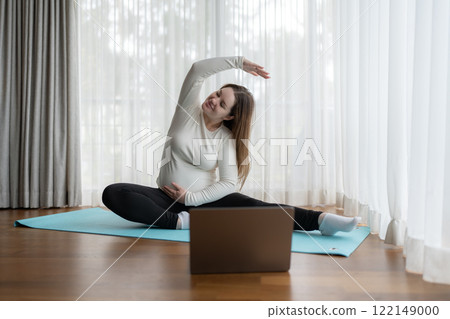 Young pregnant woman practicing yoga at home with laptop. Expectant mother doing prenatal training class.  122149000