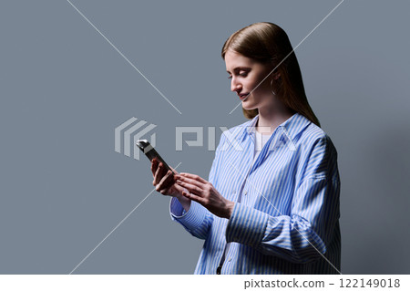 Teenage student girl in blue shirt holding smartphone in hands on grey studio background 122149018