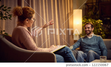Female psychologist with notes gesturing during a session with a male client, both seated in a warmly lit room with a calm atmosphere. 122150067
