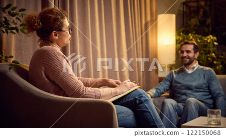 Female psychologist with notes explaining something to a male client seated on a couch in a warm, cozy therapy space. Female psychologist with notes explaining something to a male client seated on a couch in a warm, cozy therapy space. 122150068