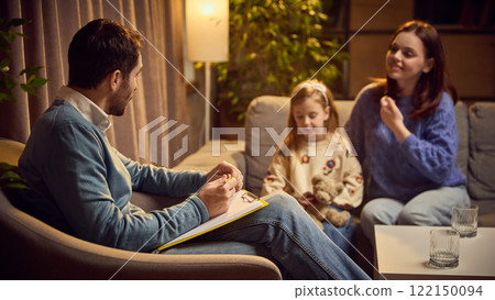 Child, girl sits next to her mother during therapy session, holding toy as the psychologist listens attentively. Supportive and nurturing setting. 122150094