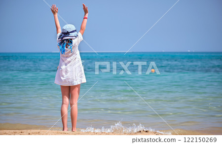 Rear view of child girl in white dress and hat standing barefoot with arms raised up on sandy beach enjoying tropical vacations looking at blue sea water 122150269