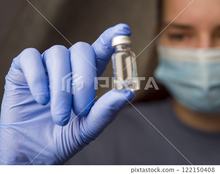 Medical professional examining clear liquid vial, wearing blue latex gloves and surgical mask in clinical setting Medical professional examining clear liquid vial, wearing blue latex gloves and surgical mask in clinical setting 122150408