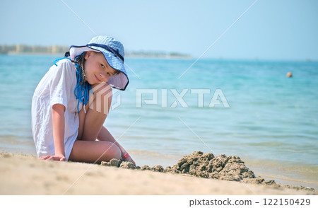 Happy hild girl in big hat and white dress playing alone with wet sand on sandy beach near clear sea lagoon water Happy hild girl in big hat and white dress playing alone with wet sand on sandy beach near clear sea lagoon water 122150429