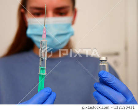 Healthcare professional wearing protective blue gloves, filling syringe with vaccine from medical vial, representing careful medical procedure in clinical environment Healthcare professional wearing protective blue gloves, filling syringe with vaccine from medical vial, representing careful medical procedure in clinical environment 122150431