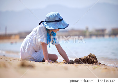 Happy hild girl in big hat and white dress playing alone with wet sand on sandy beach near clear sea lagoon water 122150432