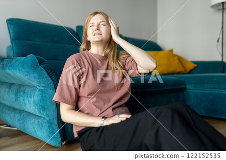 Depressed young woman sitting near couch in the living room at home, Frustrated confused female feels unhappy problem in personal life. Exhausted girl portrait suffering migraine. Hand on head. 122152535
