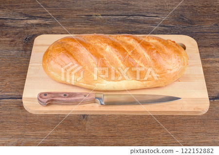 A kitchen knife and a whole loaf of white bread on a wooden cutting board. 122152882