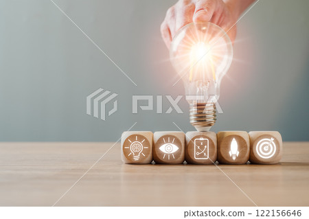 A glowing light bulb atop wooden blocks with icons, representing creativity, innovation, and strategic thinking. 122156646
