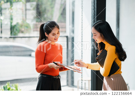 Two women in a bright office discussing work, using a digital tablet, dressed in vibrant colors. 122156664