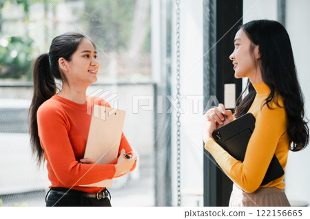 Two young women in casual attire chatting and smiling in a modern office environment, holding notebooks. 122156665