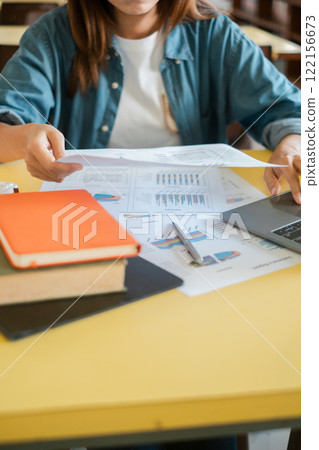 A woman reviews financial documents at a desk with a laptop, notebooks, and charts, highlighting a professional office environment. 122156673