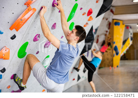 Male mountaineer climbing artificial rock wall without his belay indoors Male mountaineer climbing artificial rock wall without his belay indoors 122158471