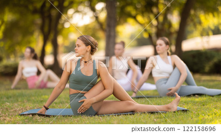 Sporty young girls performing seated twisting asana Matsyendrasana on mat in in summer park during daily yoga session Sporty young girls performing seated twisting asana Matsyendrasana on mat in in summer park during daily yoga session 122158696